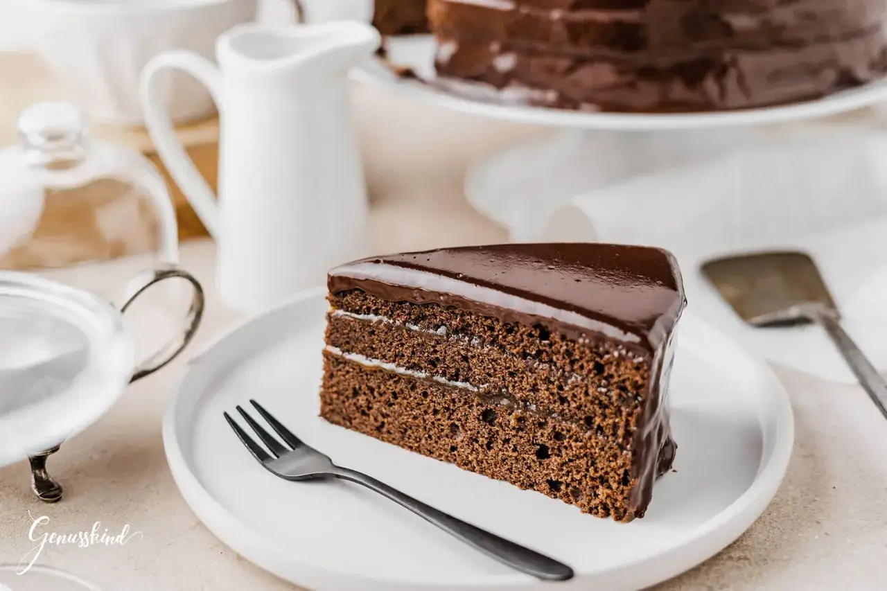 Ein Stück Sacher Torte mit glänzender Schokoladenglasur und zarter Füllung auf einem weißen Teller, bereit zum Genuss.