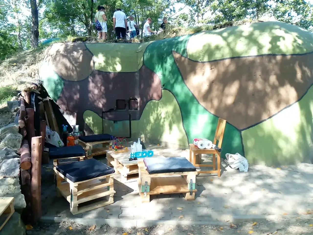 Oryginalny bunkier 44, teraz z wygodnymi siedziskami z palet i zabawkami dla dzieci, otoczony zielenią.
