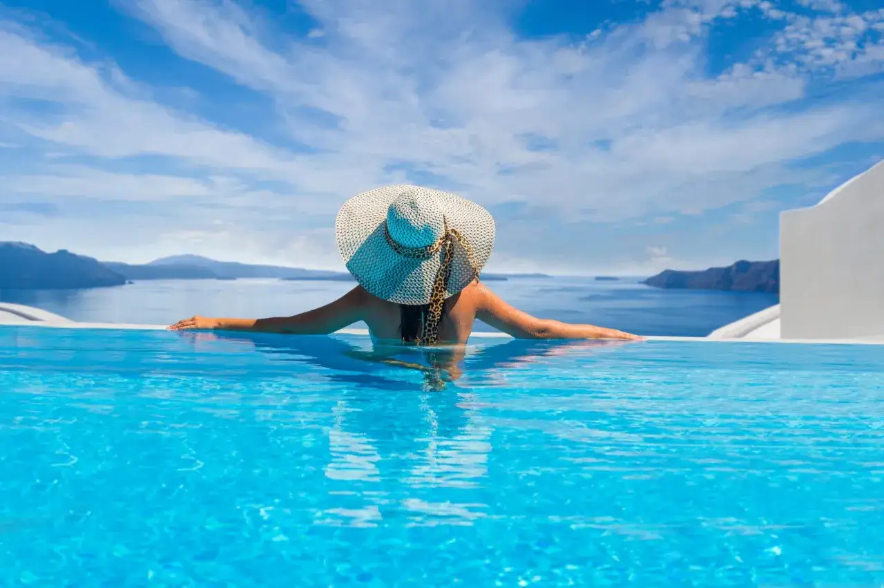 Mujer con sombrero descansa en una piscina infinita, contemplando el mar y las islas bajo un cielo azul.