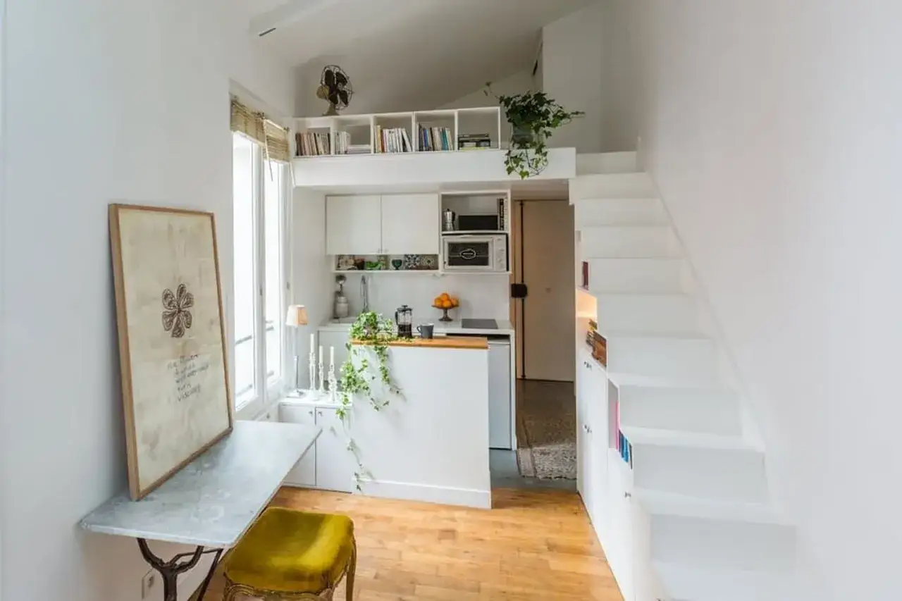 Un petit appartement optimisé avec une cuisine, un coin repas et un lit en mezzanine accessible par des escaliers blancs.