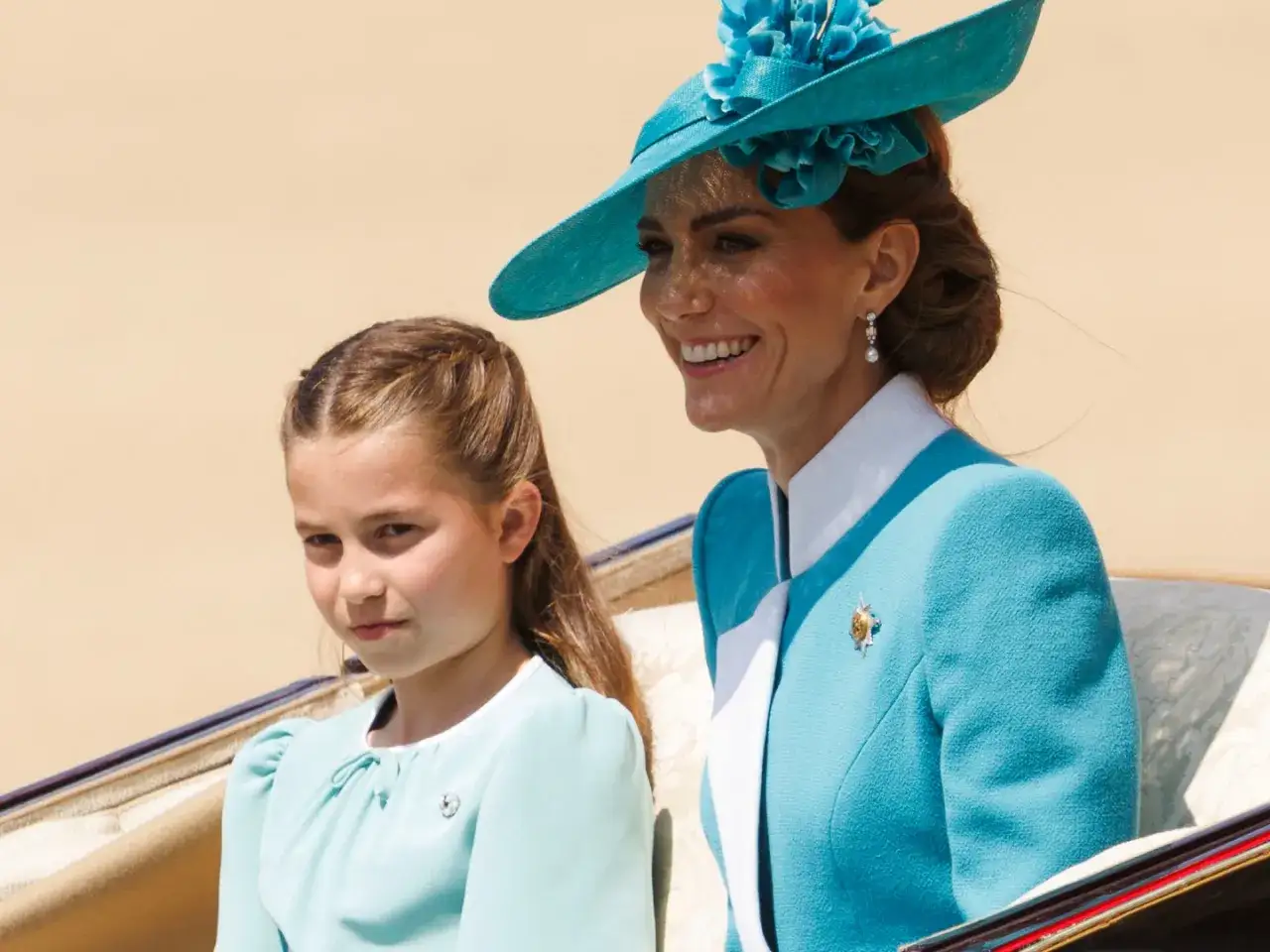 Księżniczka Charlotte w błękitnej sukience obok uśmiechniętej mamy w turkusowym kapeluszu.