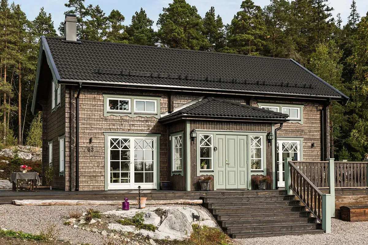 Vackert lgb timmerhus med gr&ouml;na detaljer, omgivet av skog. En inbjudande entr&eacute; med trappa leder upp till huset.