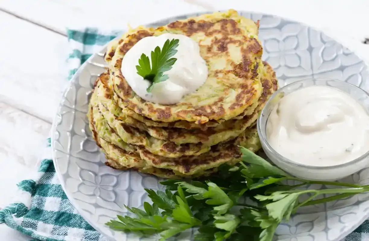 Pile de galettes de courgette dorées, garnies d'une cuillère de crème et de persil frais, servies avec une sauce crémeuse.