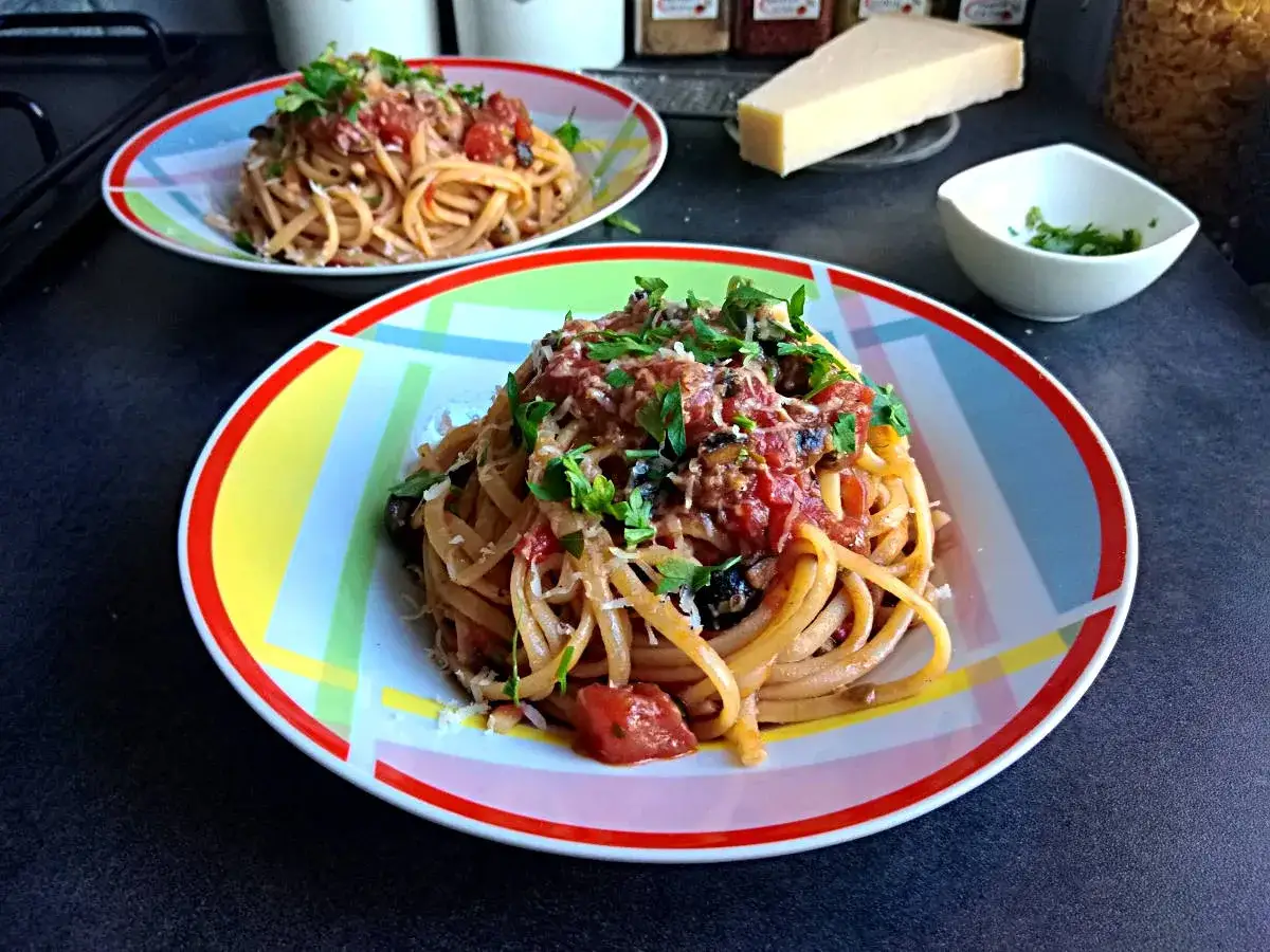 Dwa talerze spaghetti z pomidorowym sosem do makaronu, posypane serem i ziołami. Obok kawałek parmezanu i miseczka z posiekaną natką.