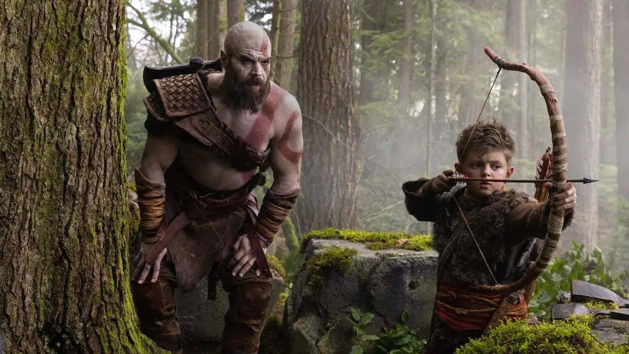 Kratos, z bliznami i tatuażami, obserwuje syna ćwiczącego łucznictwo. Scena z serialu 