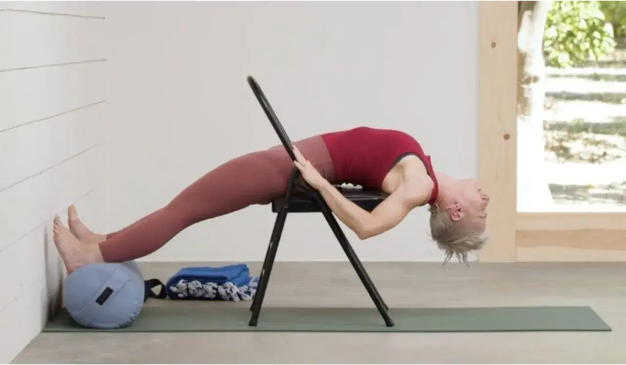 Kobieta wykonuje głęboki łuk w tył w pozycji leżącej na krześle, wspierając stopy na wałku. Praktyka iyengar yoga.