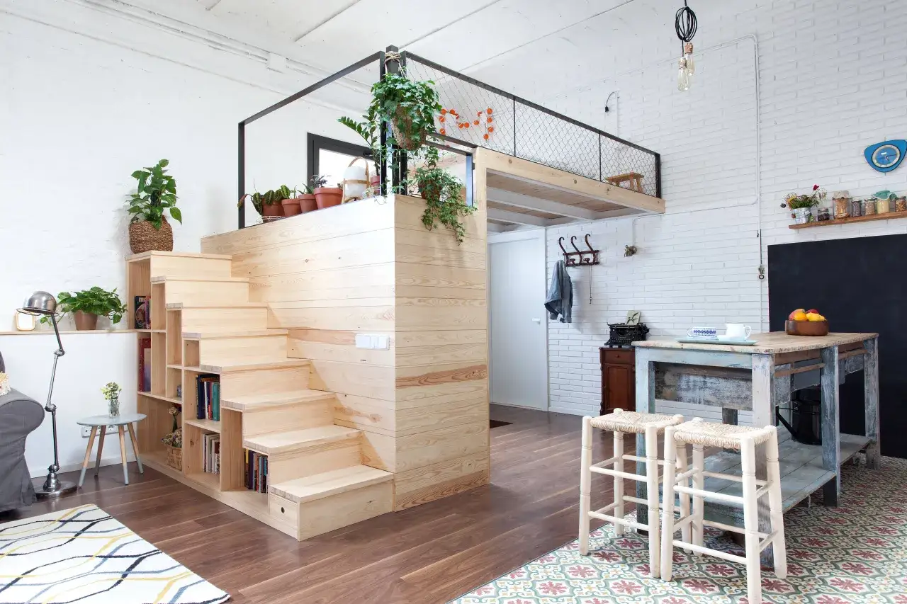Am&eacute;nagement d'un ancien garage en habitation : escalier en bois menant &agrave; une mezzanine, cuisine ouverte, et coin salon cosy.