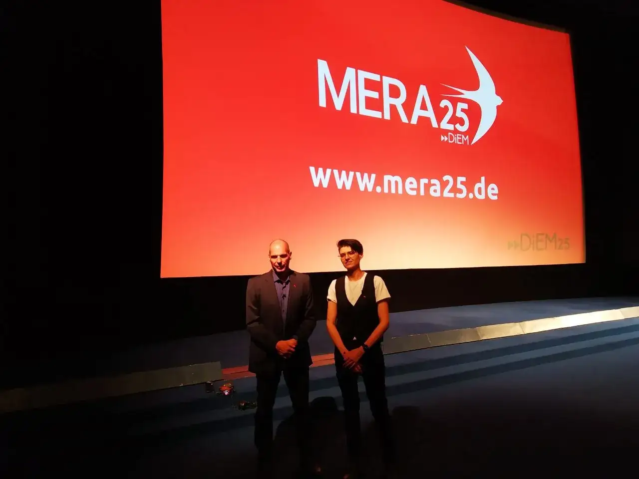 Zwei Männer stehen vor einer roten Leinwand mit dem Logo von MERA25. Die Leinwand zeigt die Website www.mera25.de. Die Kritik an Diem25 wird hier vielleicht thematisiert.