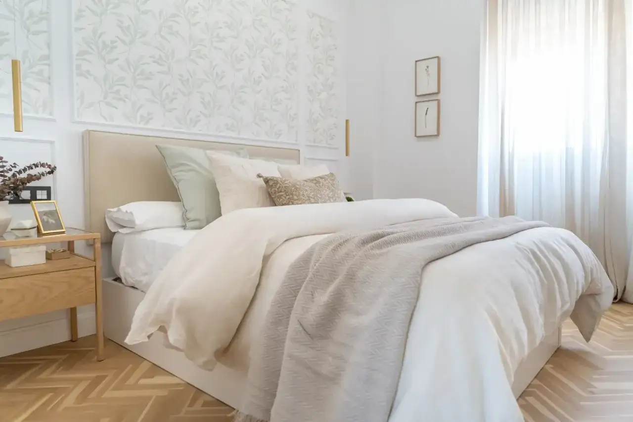 Habitaciones con papel pintado de hojas verdes y paredes pintadas en blanco. Cama con ropa de cama blanca y manta gris.