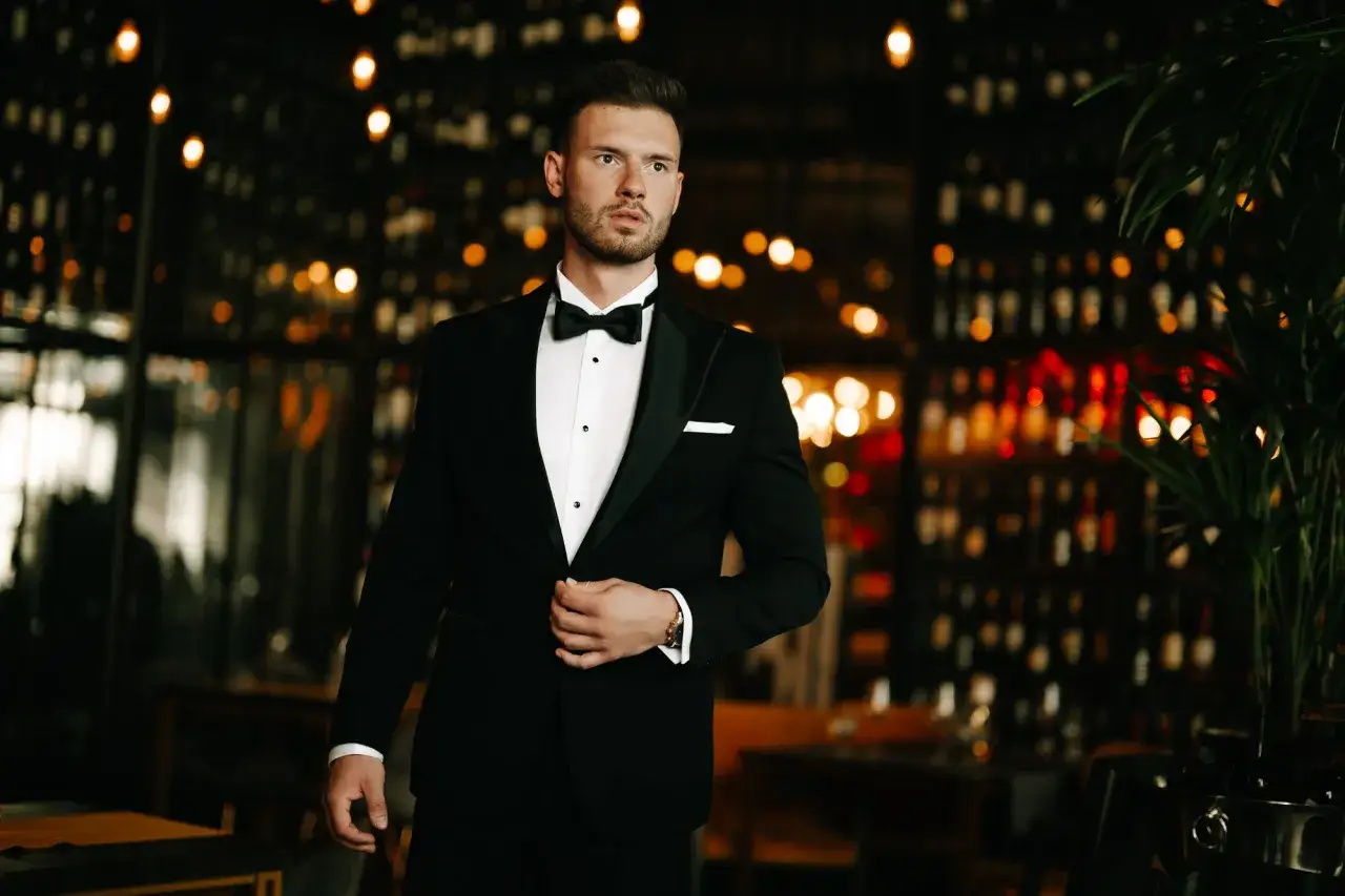 Elegancki mężczyzna w czarnym smokingu, z muszką i białą poszetką, stoi na tle rozświetlonego wnętrza.