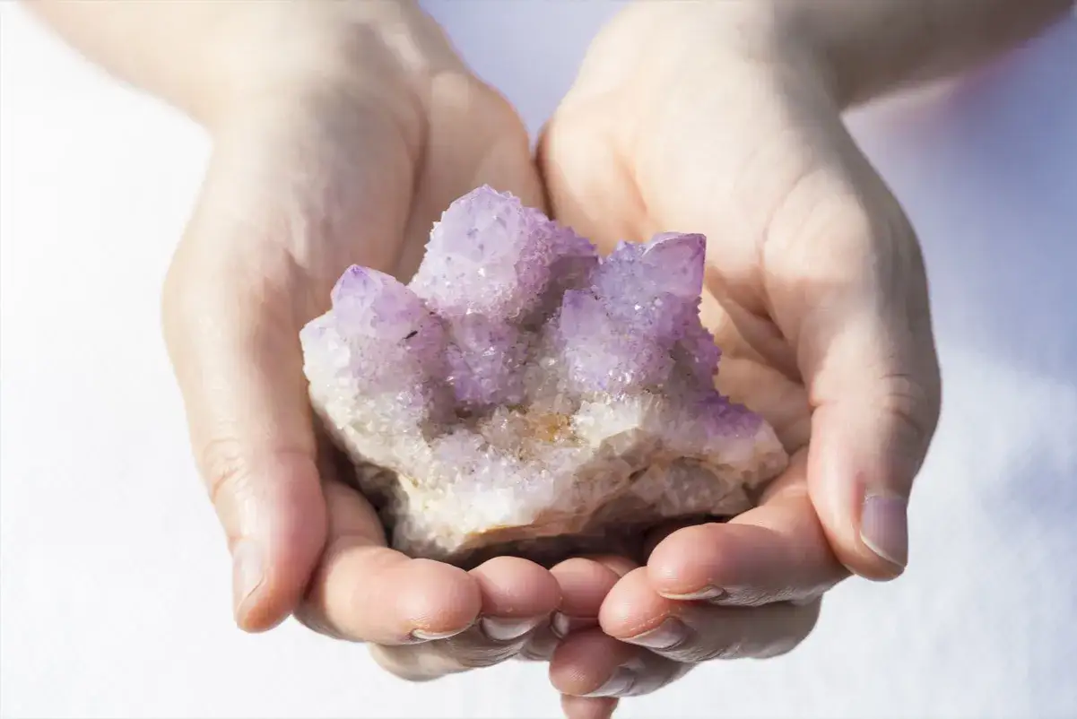 Manos sostienen un racimo de cristales energ&eacute;ticos de amatista p&uacute;rpura y blanco, brillando con luz.