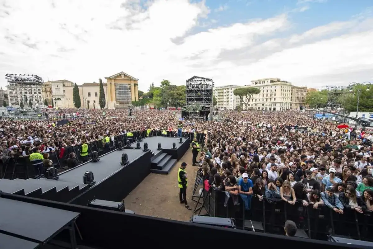 Un'enorme folla attende gli artisti primo maggio in piazza, con un palco imponente e palazzi storici sullo sfondo.