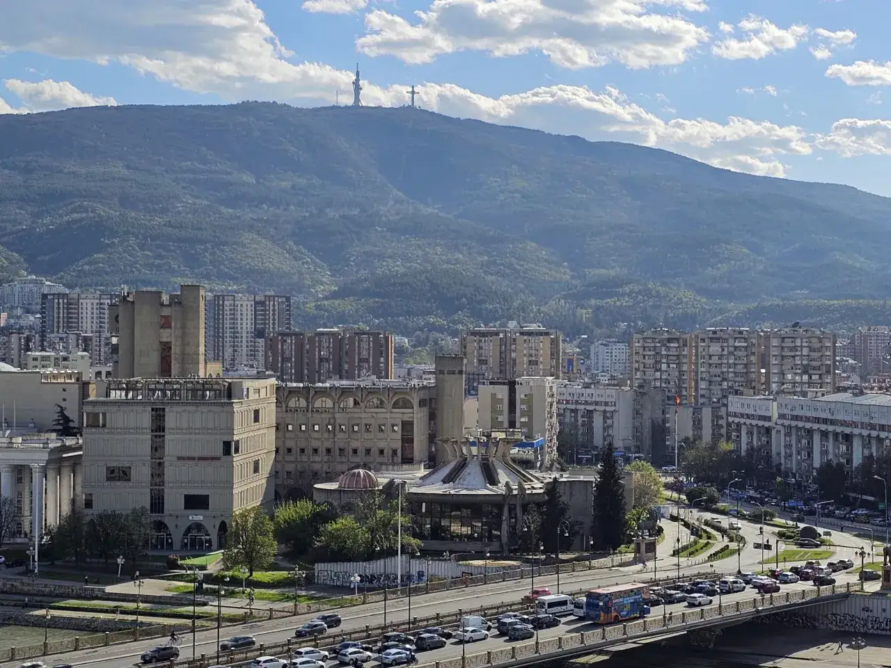 Widok na Skopje, stolicę Macedonii P&oacute;łnocnej, z g&oacute;rami i krzyżem na wzg&oacute;rzu w tle.