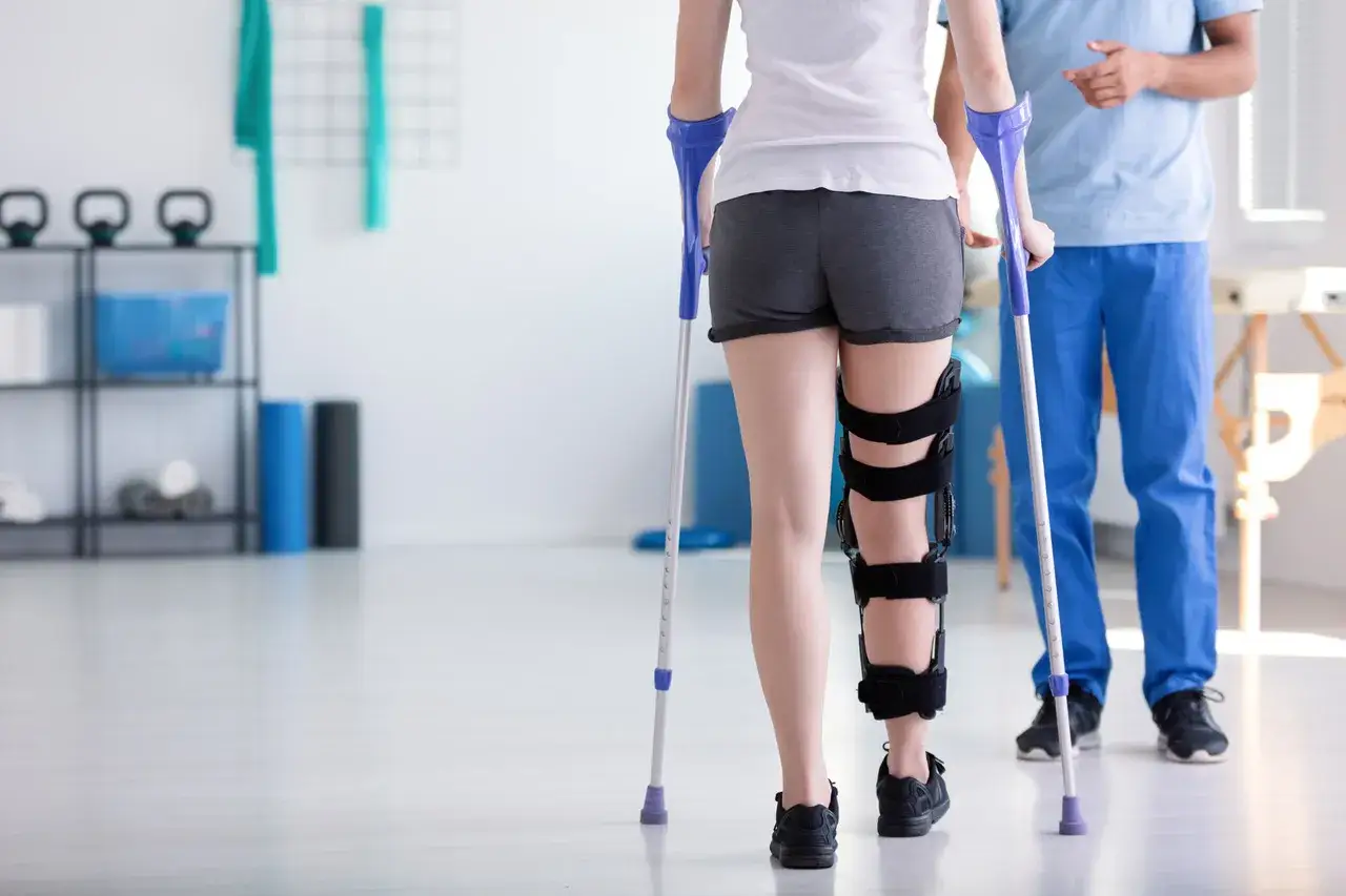 Fizjoterapeuta uczy, jak chodzić o kulach, wspierając pacjentkę z ortezą na nodze w gabinecie rehabilitacyjnym.