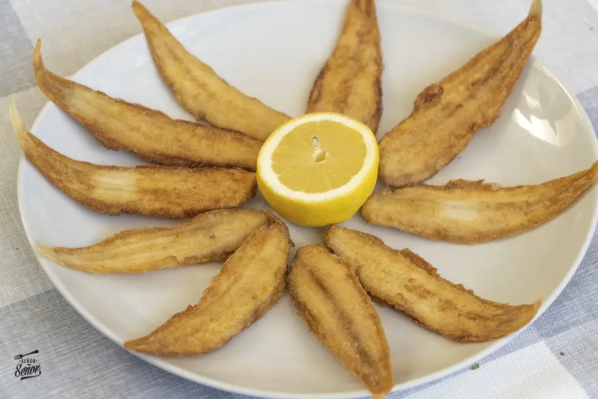 Delicioso acedia pescado frito, crujiente y dorado, servido en un plato blanco con una rodaja de limón en el centro.