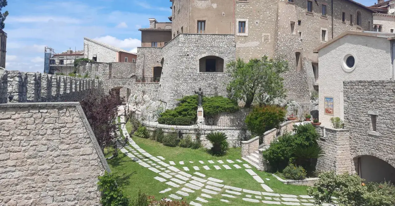 Un giardino con un sentiero di pietre conduce a una statua nel centro storico di Rieti. Edifici in pietra e vegetazione rigogliosa creano un'atmosfera suggestiva.