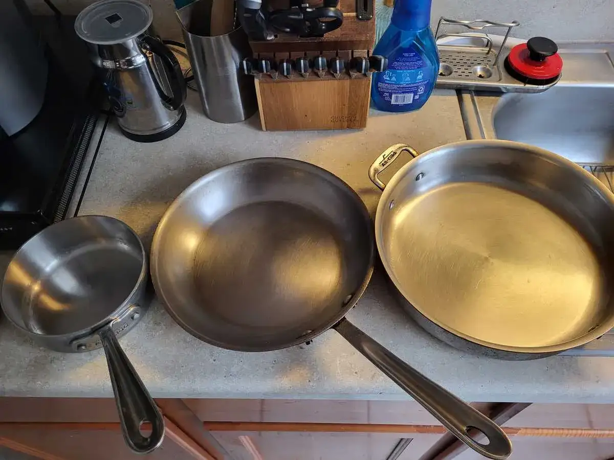 Tre stekpannor på en bänk: en liten i rostfritt stål, en större kolstål och en stor rostfri. Valet mellan kolstål vs rostfritt stål stekpanna beror på matlagningen.