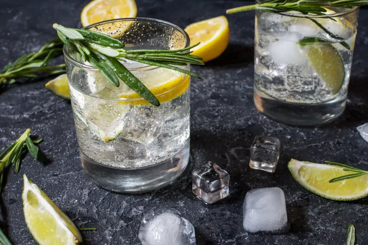 Orzeźwiający gin z tonikiem z plasterkami cytryny, limonki i gałązkami świeżego rozmarynu. Widać kostki lodu i pęcherzyki w napoju, co sugeruje jego orzeźwiające skutki.