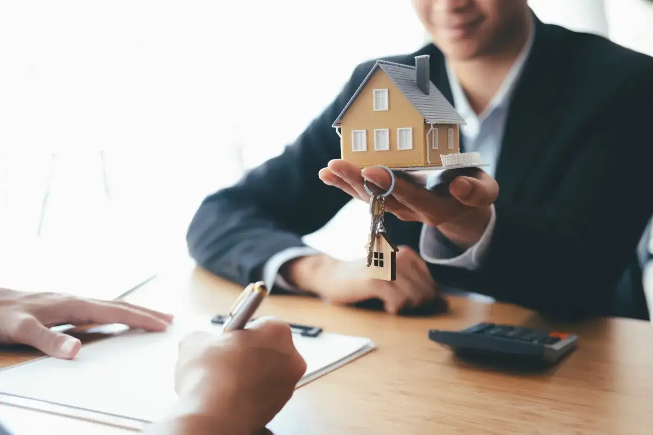 Un homme d'affaires présente une maquette de maison et des clés, symbolisant un prêt immobilier sans apport nouvelle loi. Une personne signe un document.