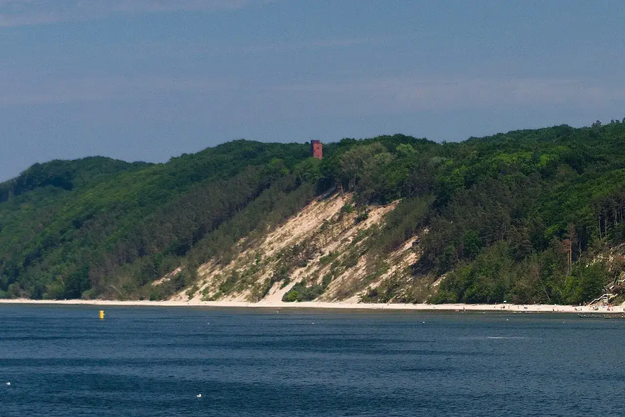 Zielone wzgórza z piaszczystym zboczem, na szczycie którego stoi ceglana wieża. Widok na morze i plażę. Biała góra w oddali.