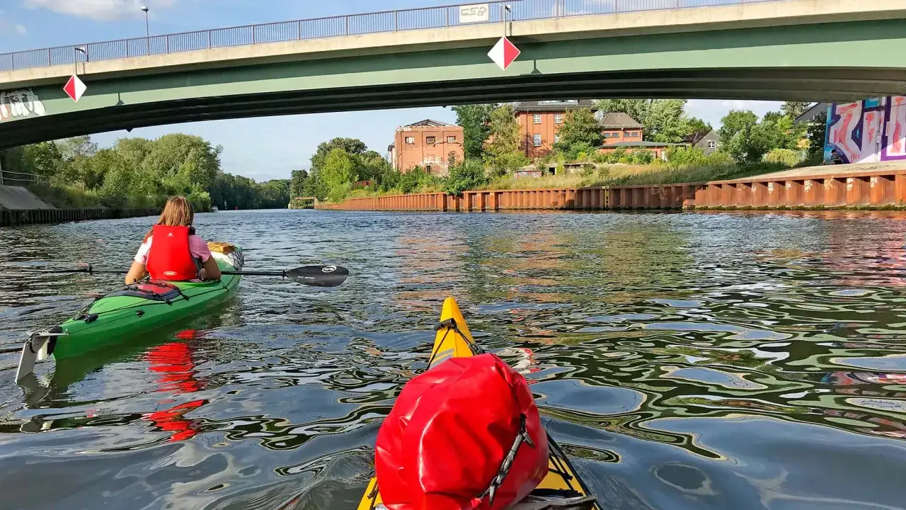 Zwei Kajaks gleiten unter einer Br&uuml;cke hindurch. Eine Person im gr&uuml;nen Boot, die andere im gelben. Ein entspanntes Paddeln in Berlin.