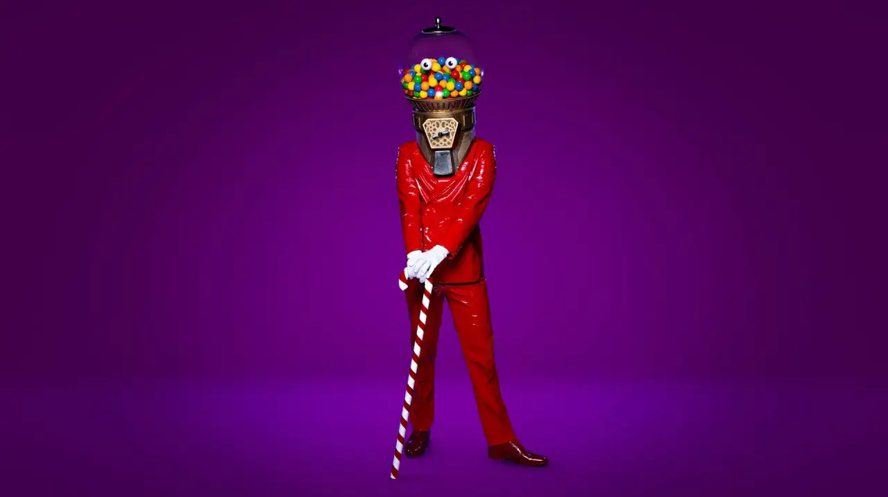 En person i röd, glansig dräkt med ett godisautomathuvud fullt av kulor. Håller en polkagrisstav. En riktig Masked Singer-tid!