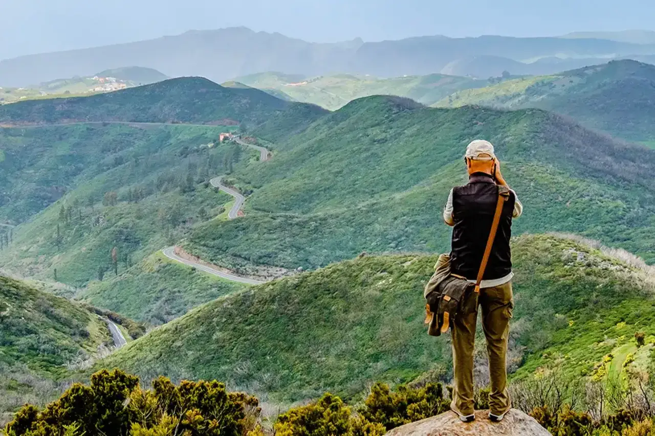 Mann mit Kamera auf Felsen, genie&szlig;t die Aussicht auf gr&uuml;ne H&uuml;gel und Serpentinenstra&szlig;en. Perfekt f&uuml;r das Wandern im November.