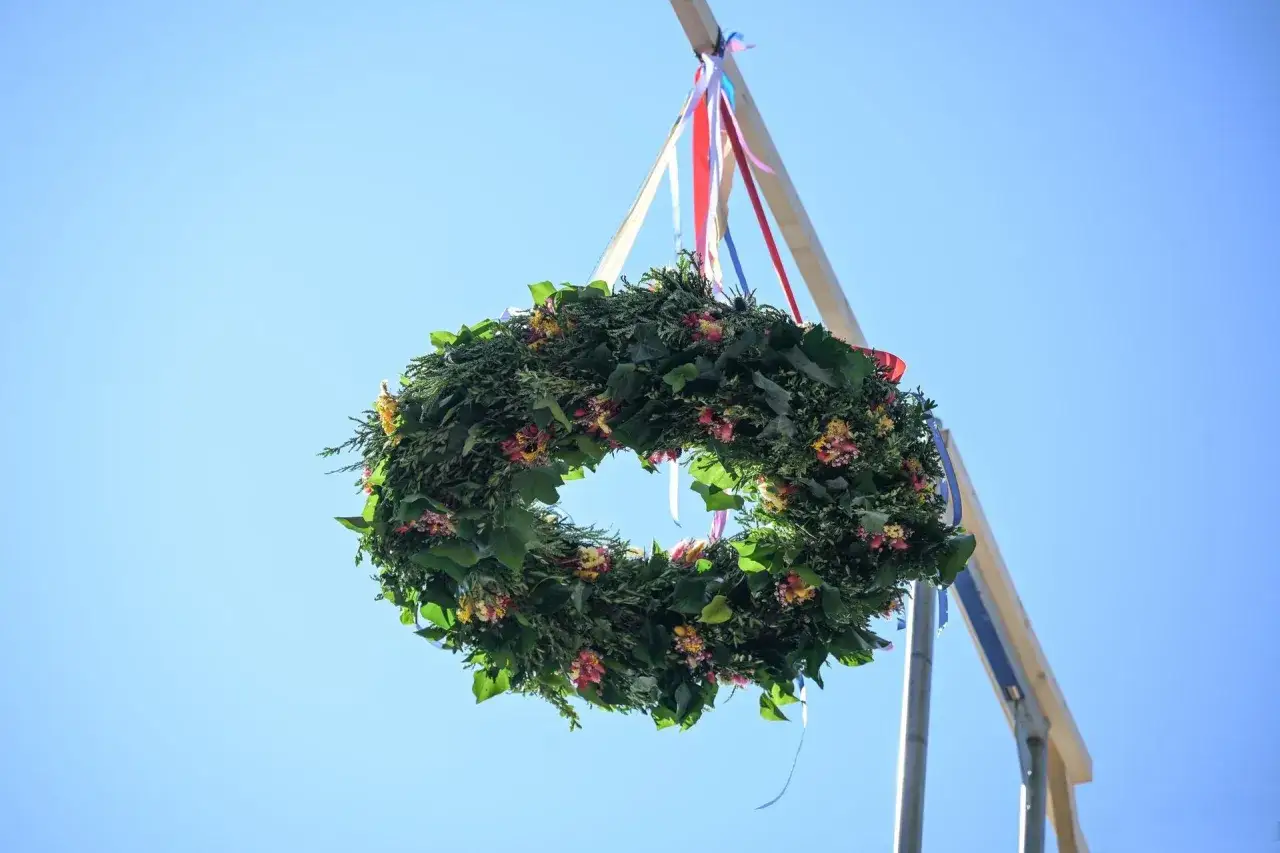 Wieniec z zieleni i kwiatów, ozdobiony kolorowymi wstążkami, wisi na drewnianym słupie. To piękna wiecha, symbol lata.