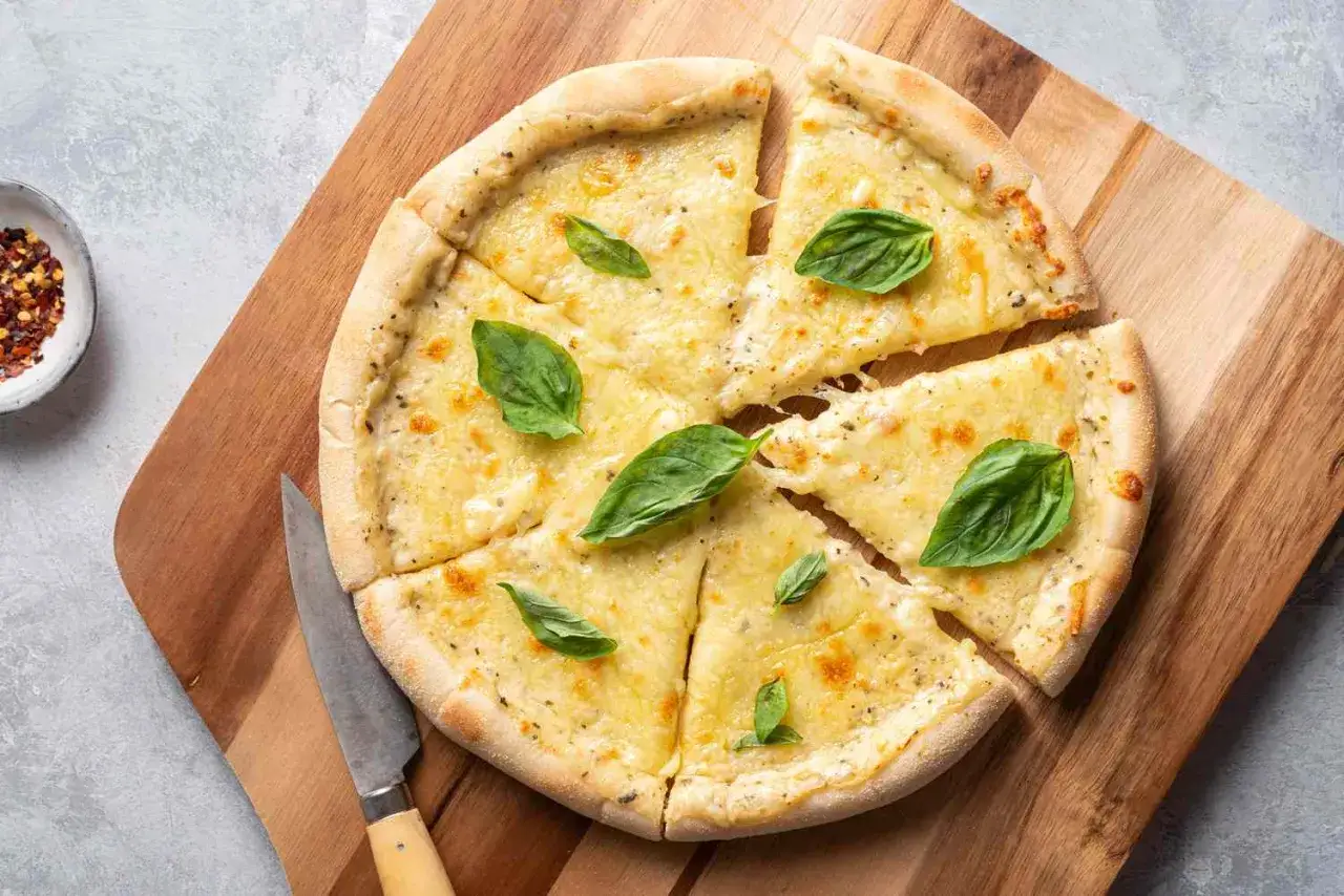 Pizza z serem i bazylią, polana kremowym, białym sosem do pizzy. Obok leży nóż i miseczka z płatkami chili.