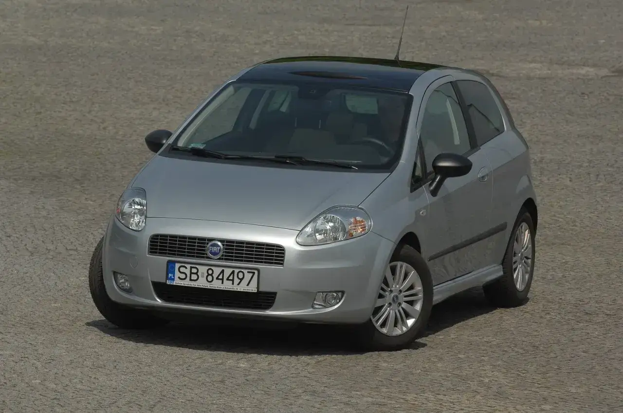 Srebrny Fiat Grande Punto z czarnym dachem i felgami. Jego żarówki są gotowe do rozświetlenia drogi.