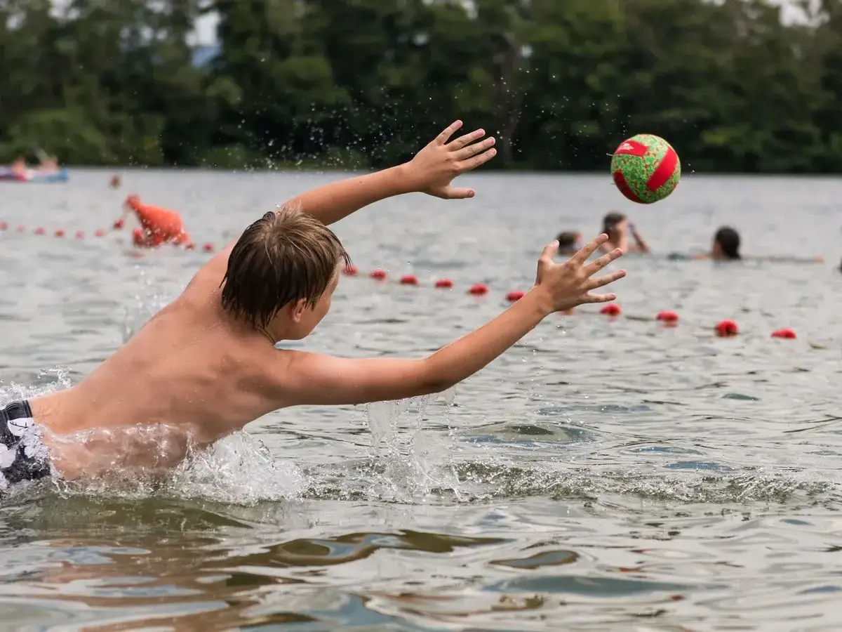 Junge versucht am Badesee Frankfurt, einen Ball zu fangen. Wasser spritzt, andere baden im Hintergrund.
