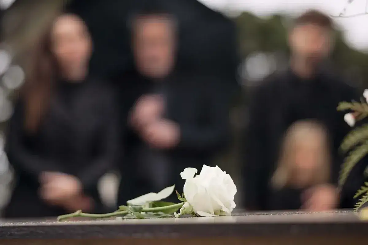 Biała róża na kamiennym nagrobku, w tle zamazane sylwetki żałobników. To obraz, który skłania do refleksji, ile trwa organizacja pogrzebu.