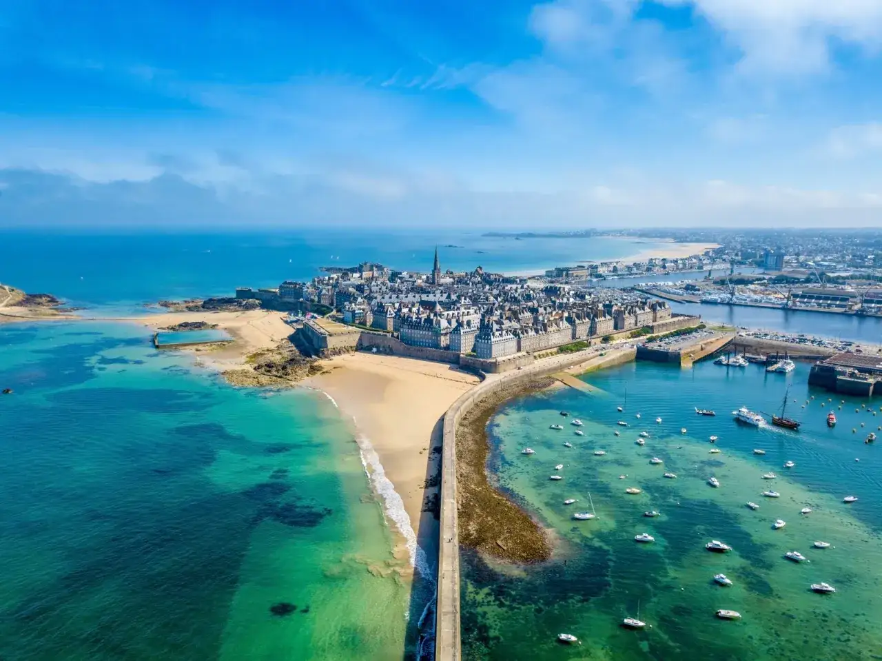 Luftaufnahme der befestigten Stadt Saint-Malo mit ihren historischen Geb&auml;uden, umgeben von t&uuml;rkisfarbenem Wasser und einem Sandstrand.