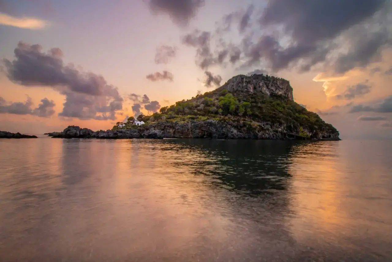 Alba sulle isole calabresi: un'isola rocciosa con case bianche si specchia nel mare calmo, sotto un cielo dai colori pastello.