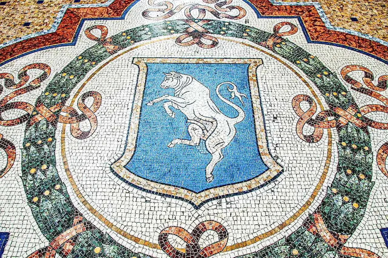 Mozaika z bykiem na niebieskiej tarczy w Galerii Emanuela w Mediolanie. Wzory i kolory tworzą bogaty dywan.