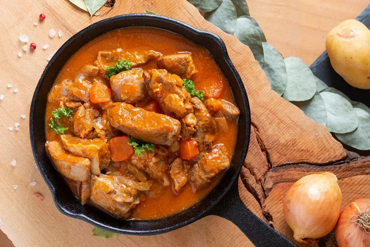 Un savoureux veau marengo mijote dans une poêle en fonte, garni de persil frais et de carottes. Des oignons et une pomme de terre complètent la scène.