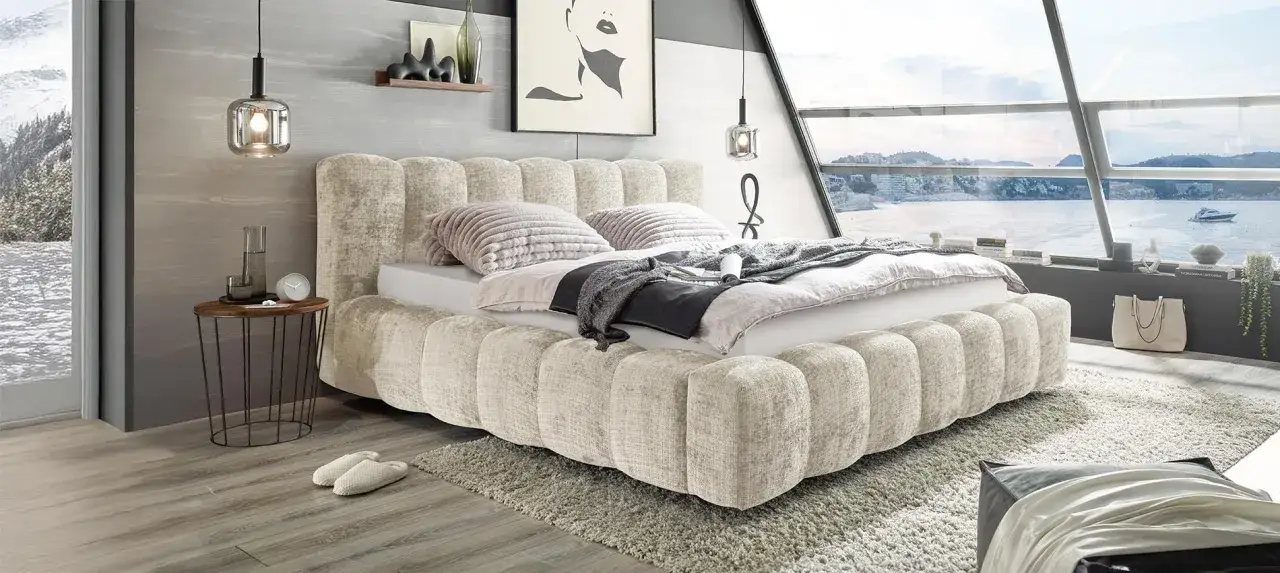 Ein luxuriöses Bett mit gepolstertem Kopfteil und Sockel, das sich fragt: welches Bett passt zu mir? Gemütlich mit Kissen und Decken, neben einem Fenster mit Meerblick.