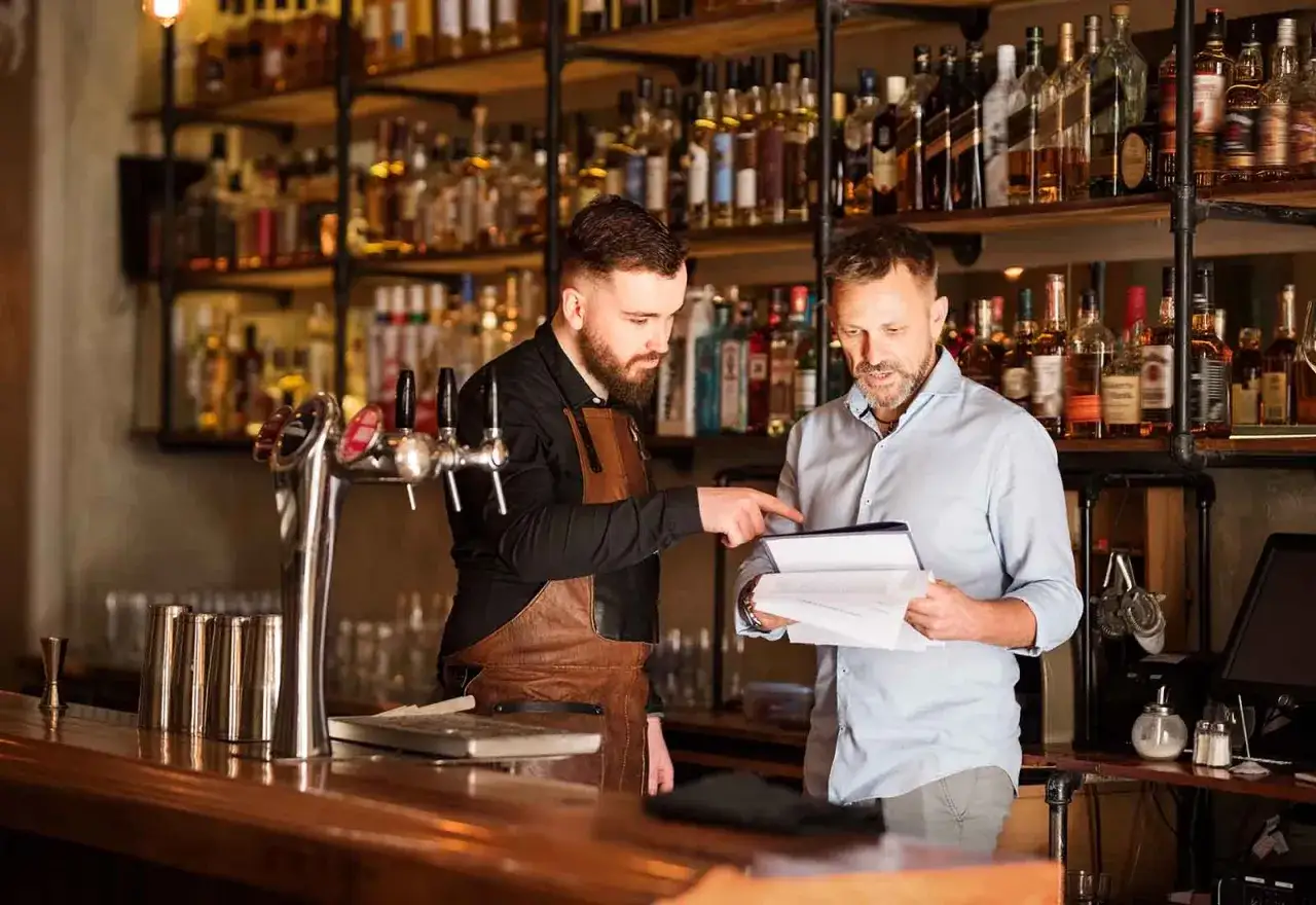 Dos hombres discuten un menú en una barra, mostrando la diferencia entre bar y restaurante. Estantes llenos de botellas de licor detrás.