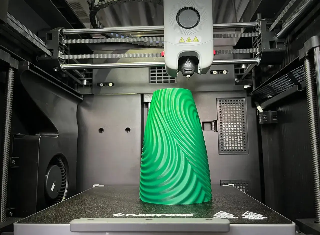 A green, wavy vase is being printed by a high resolution 3d printer.