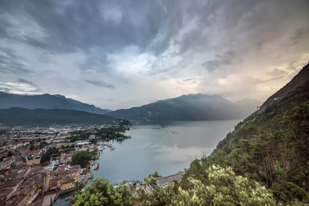Panoramica delle attrazioni di Riva del Garda con il lago e le montagne sotto un cielo nuvoloso.