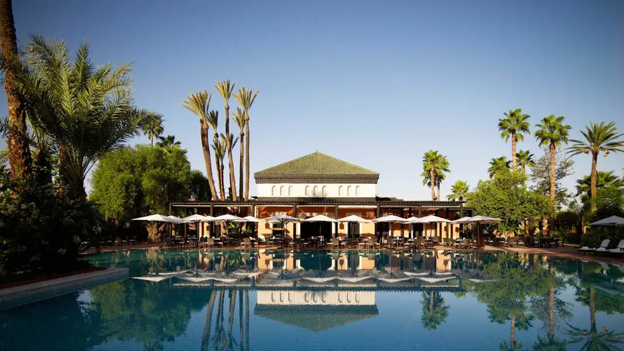Ein glitzernder Pool spiegelt die Palmen und das elegante Gebäude des La Mamounia wider, umgeben von üppiger Vegetation.