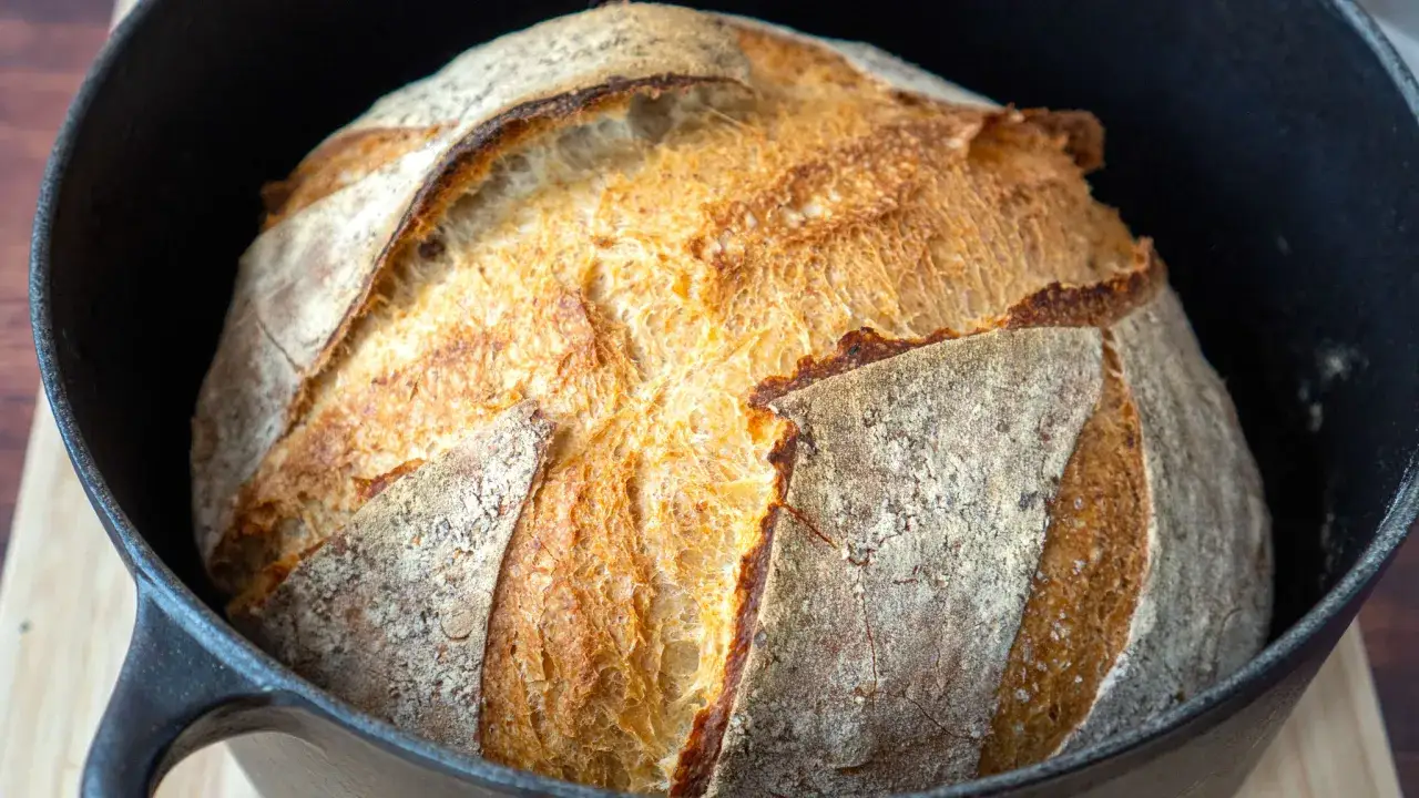 Ein frisch gebackenes, goldbraunes Brot mit knuspriger Kruste ruht in einem schwarzen Gusseisentopf. Perfekt f&uuml;r einfaches Brot backen.