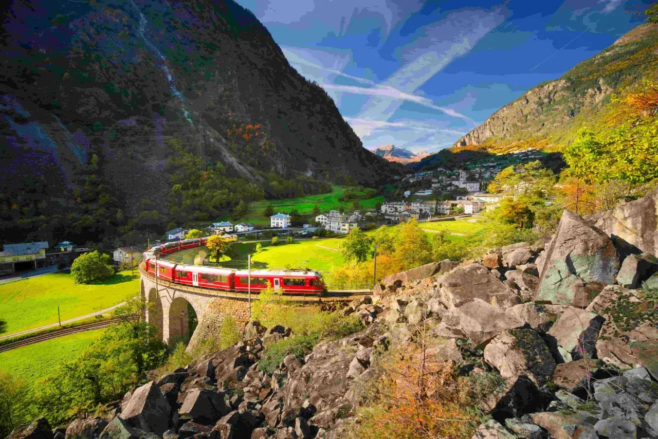 Roter Zug fährt über eine Steinbrücke in einem Tal mit Häusern und Bergen. Die Landschaft erinnert an das Eisengebirge in Frankreich.