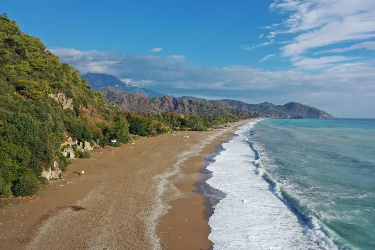 Ein einsamer antalya strand mit sanften Wellen, umgeben von gr&uuml;nen H&uuml;geln und Bergen unter blauem Himmel.