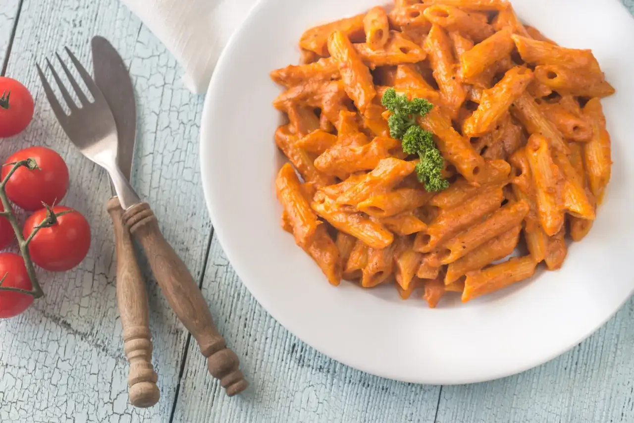 Piatto di penne alla vodka cremose, guarnite con prezzemolo, su un tavolo di legno azzurro. Accanto, pomodorini freschi e posate rustiche.