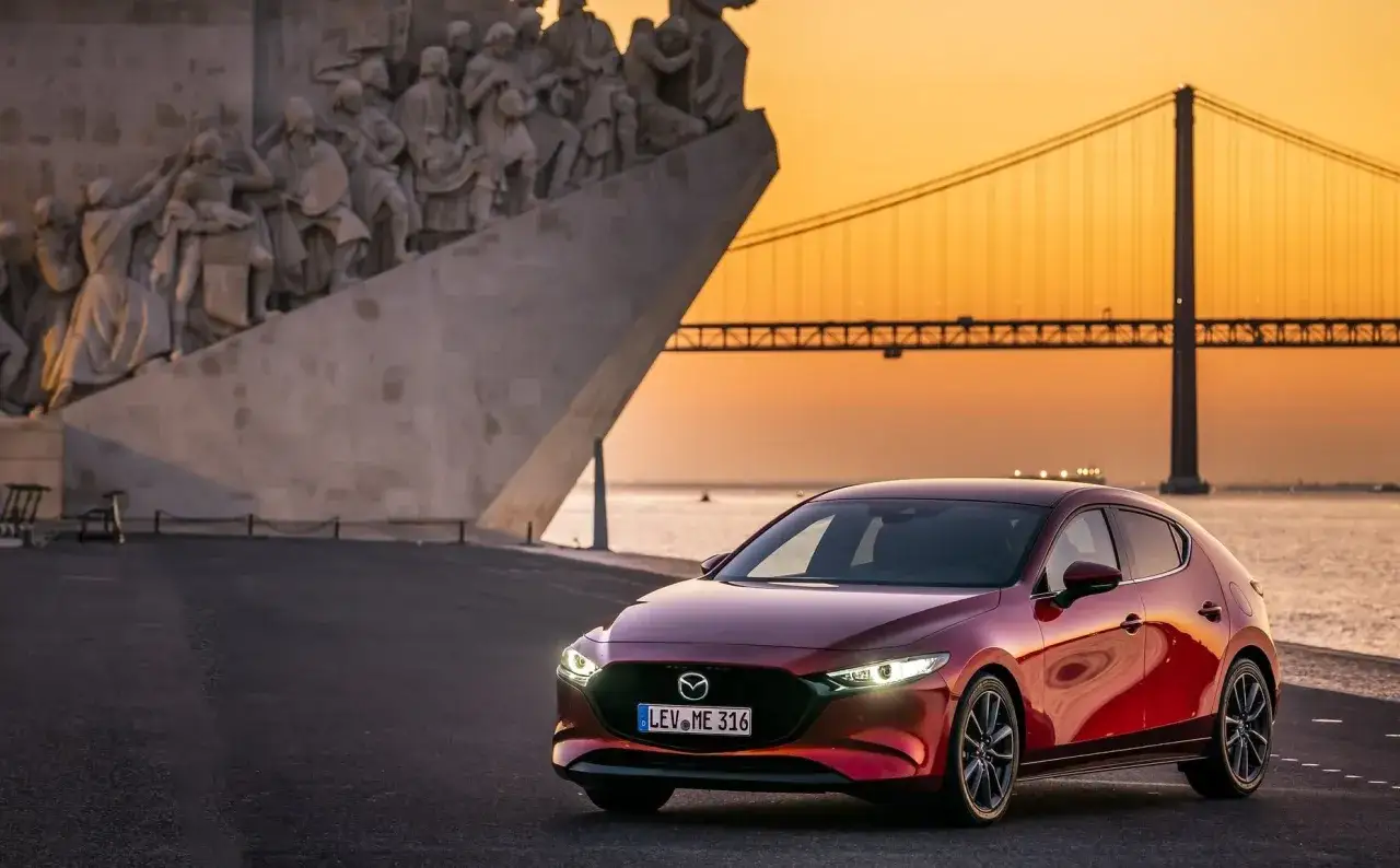 Czerwona Mazda 3, jeden z najlepszych aut, na tle pomnika i mostu o zachodzie słońca.