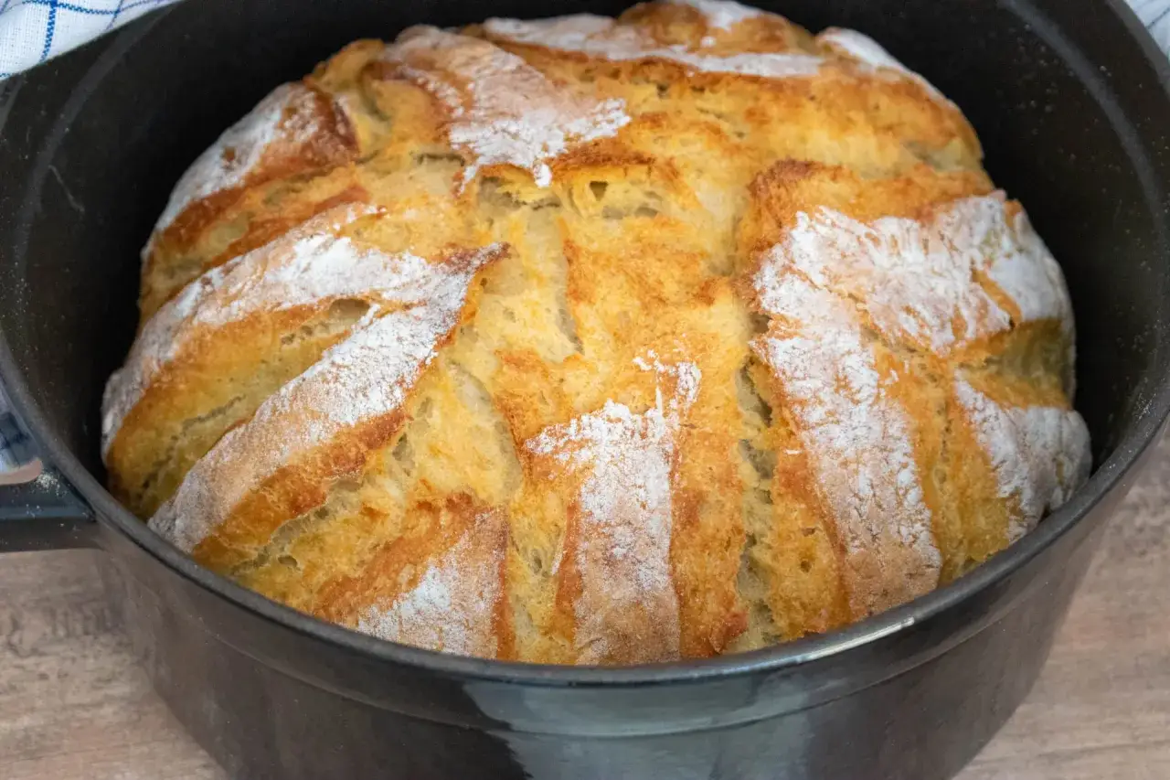 Ein frisch gebackenes, goldbraunes Kartoffelbrot mit Sauerteig ruht in einem schwarzen Gusseisentopf. Die Kruste ist mit Mehl bestäubt und zeigt tiefe Einschnitte.