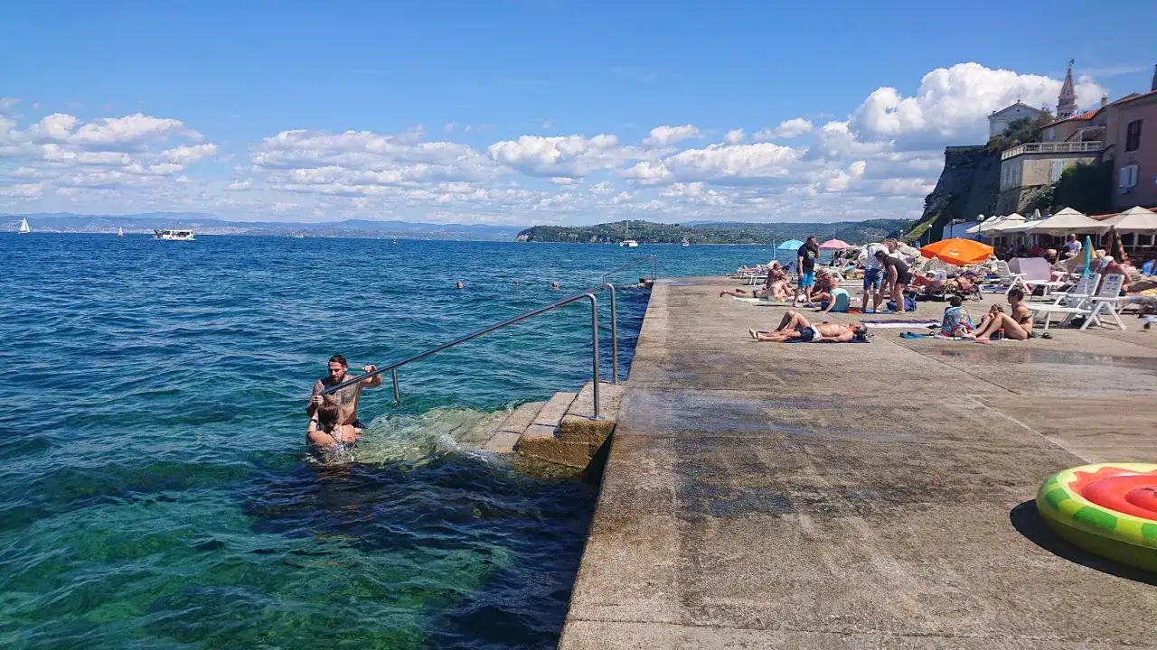 Plaża w Piran, Słowenia. Ludzie kąpią się w krystalicznie czystej wodzie, inni opalają się na betonowym molo.