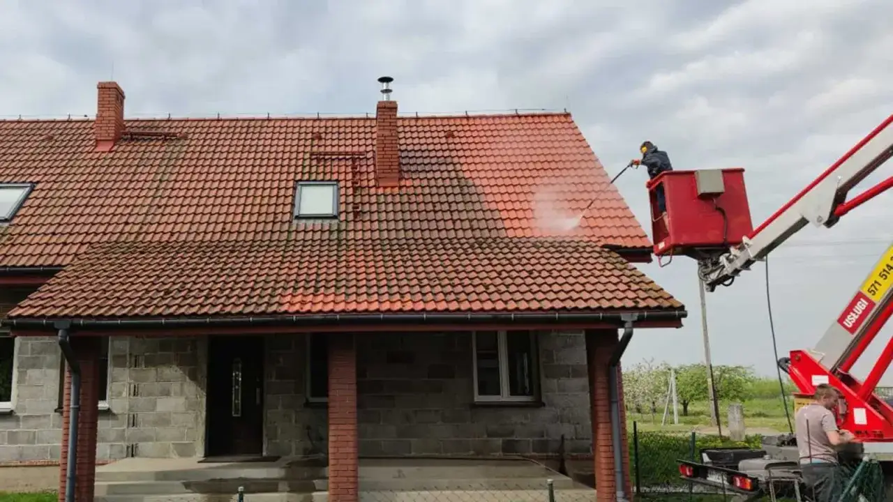 Czyszczenie dachu z użyciem podnośnika. Woda pod ciśnieniem usuwa brud z dachówki, a pracownik w koszu steruje strumieniem. Rzygownik na kominie jest już czysty.