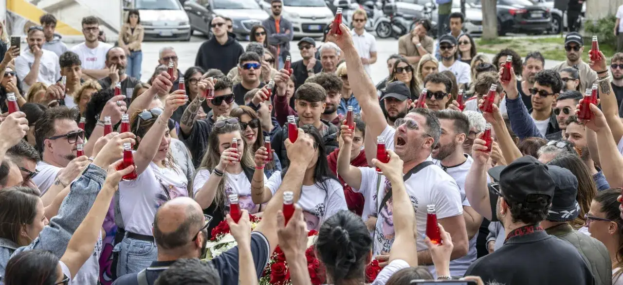 Un gruppo di persone alza bottigliette rosse in un brindisi, forse una canzone per salutare colleghi, con fiori rossi in primo piano.